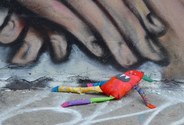 Bo the little rainbow coloured stuffed monster is lying on the concrete sidewalk at the edge of a building. On the building is a street art painting of two hands with their fingers pointed downwards. Bo is lying under the fingers. 