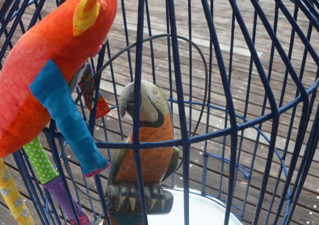 Bo, the stuffed rainbow coloured monster is looking into a metal cage where there is a wooden parrot on a perch. 