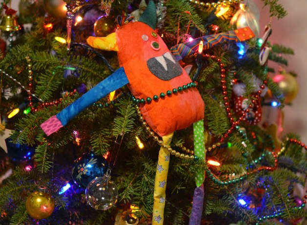 bo, the rainbow coloured stuffed monster is tied to the Christmas tree with sparking green, gold, and red beaded cord 