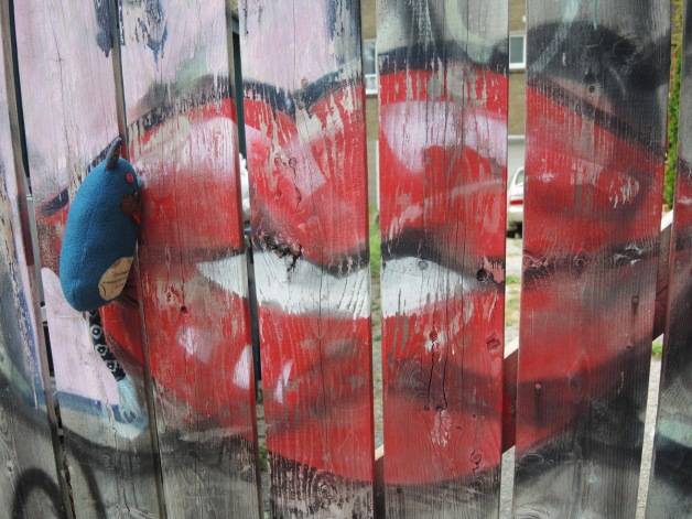 Edgar the little blue stuffed monster is perched on a wood fence with his tongue out beside a big set of red lips painted on the fence, graffiti in the alley 